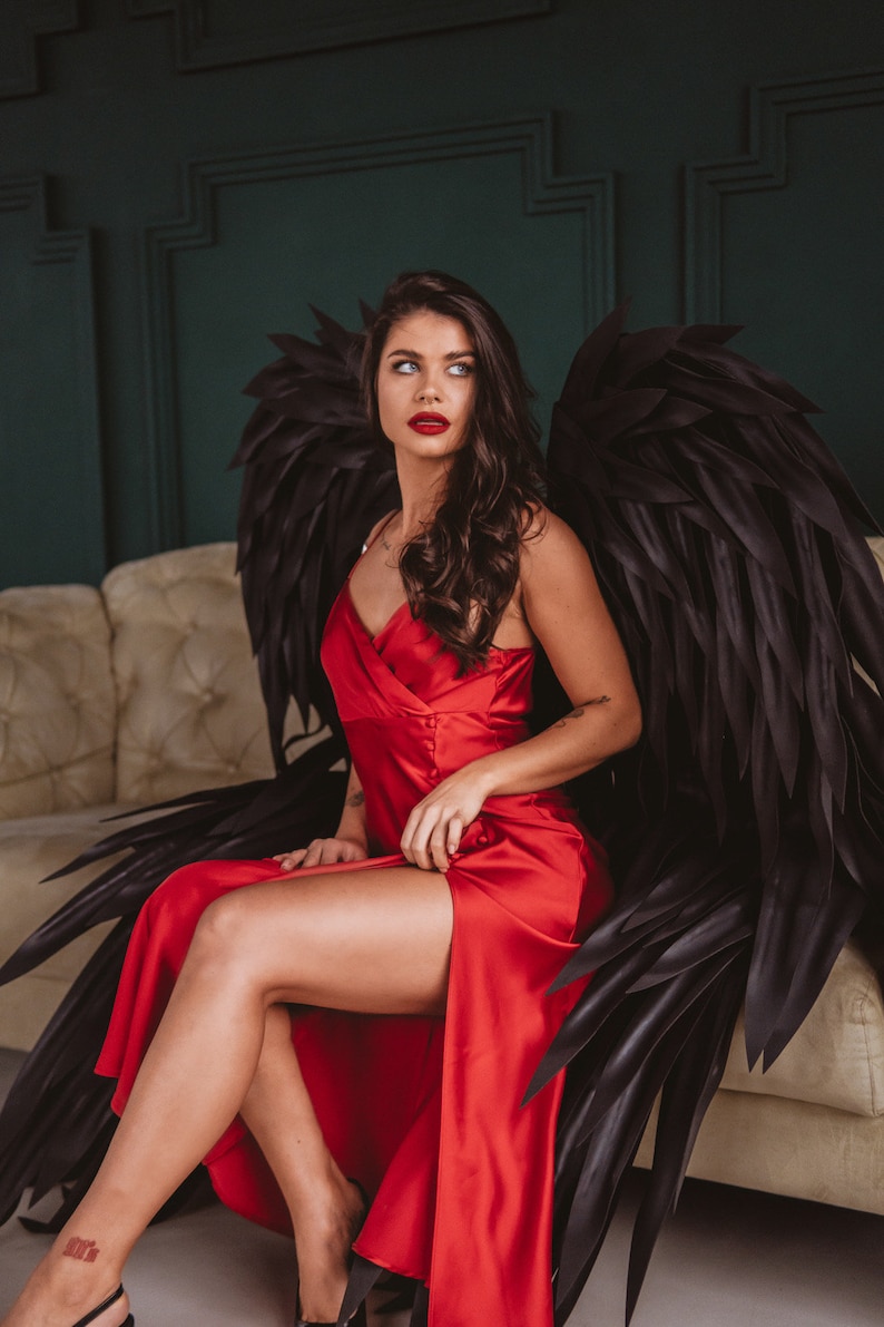 May include: A woman wearing a red satin dress and large black feathered angel wings, seated on a beige sofa. The dress has a high slit, and the wings are spread behind her. The background is a dark green wall.