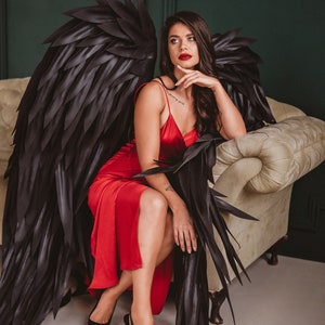 May include: A woman wearing a red satin dress and black high heels, seated on a cream-colored sofa. Large black angel wings are behind her. The background is a dark green wall.