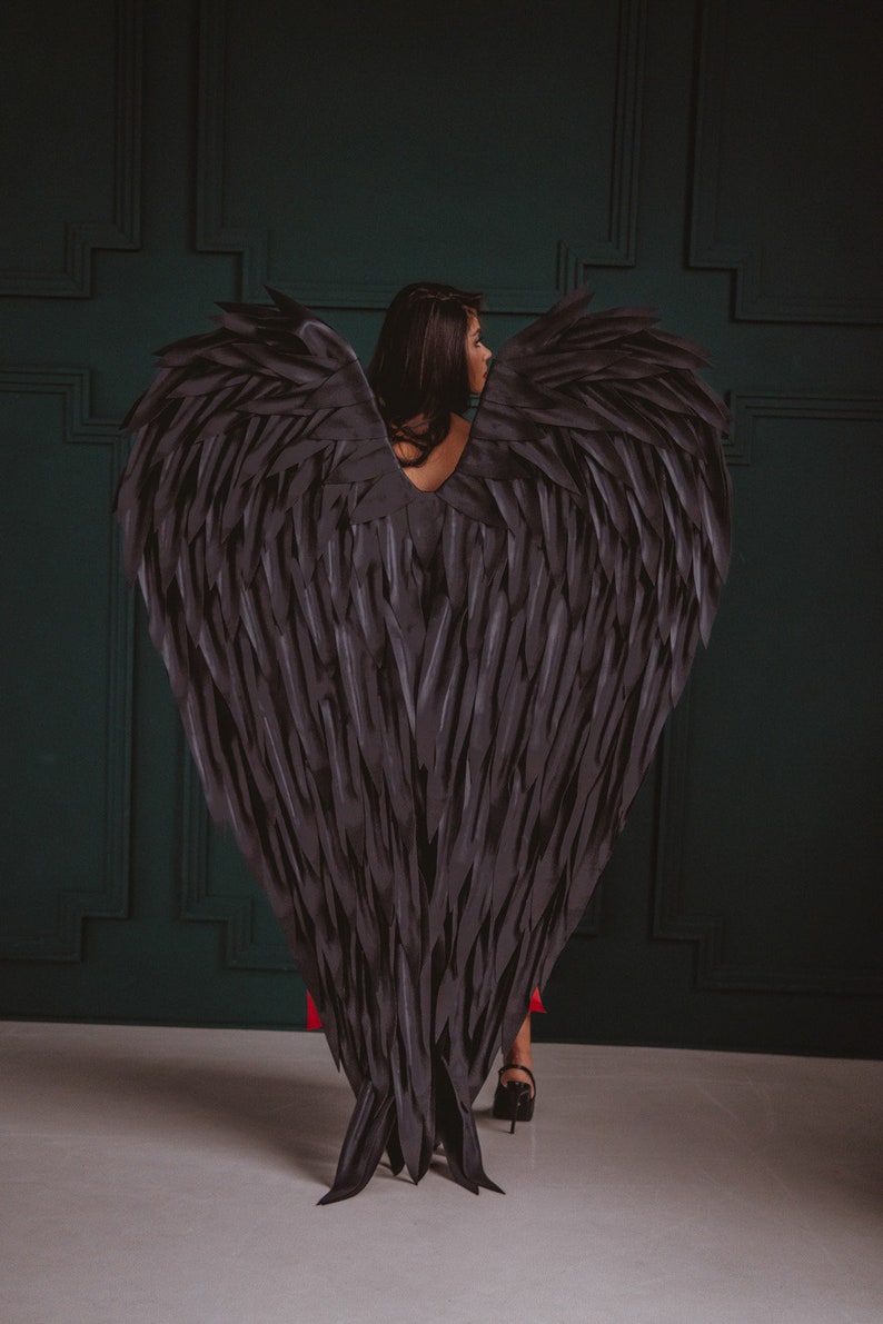 May include: A person wearing large, black angel wings. The wings are made of layered, textured material, creating a feathered appearance. The person is facing away from the camera, with dark hair and a red dress visible.