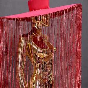 Puede incluir: Maniquí dorado con un sombrero grande de color rosa brillante con ala ancha. El sombrero está adornado con un ribete brillante. Largas y brillantes flecos rojos cuelgan del sombrero, creando un efecto de cortina. El maniquí está sobre un fondo gris.