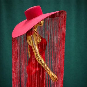 Könnte beinhalten: Ein roter Hut mit breiter Krempe und zylindrischer Krone, verziert mit einem Fransenbehang aus roten Perlen. Der Hut ist Teil eines roten Kleiderensembles auf einer goldenen Schaufensterpuppe, vor einem dunkelgrünen Hintergrund. Das Design des Hutes ist auffällig und elegant.