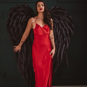 May include: A woman in a red satin dress with black angel wings. The dress has a button closure and spaghetti straps. She is wearing black heels. The background is a dark green wall.