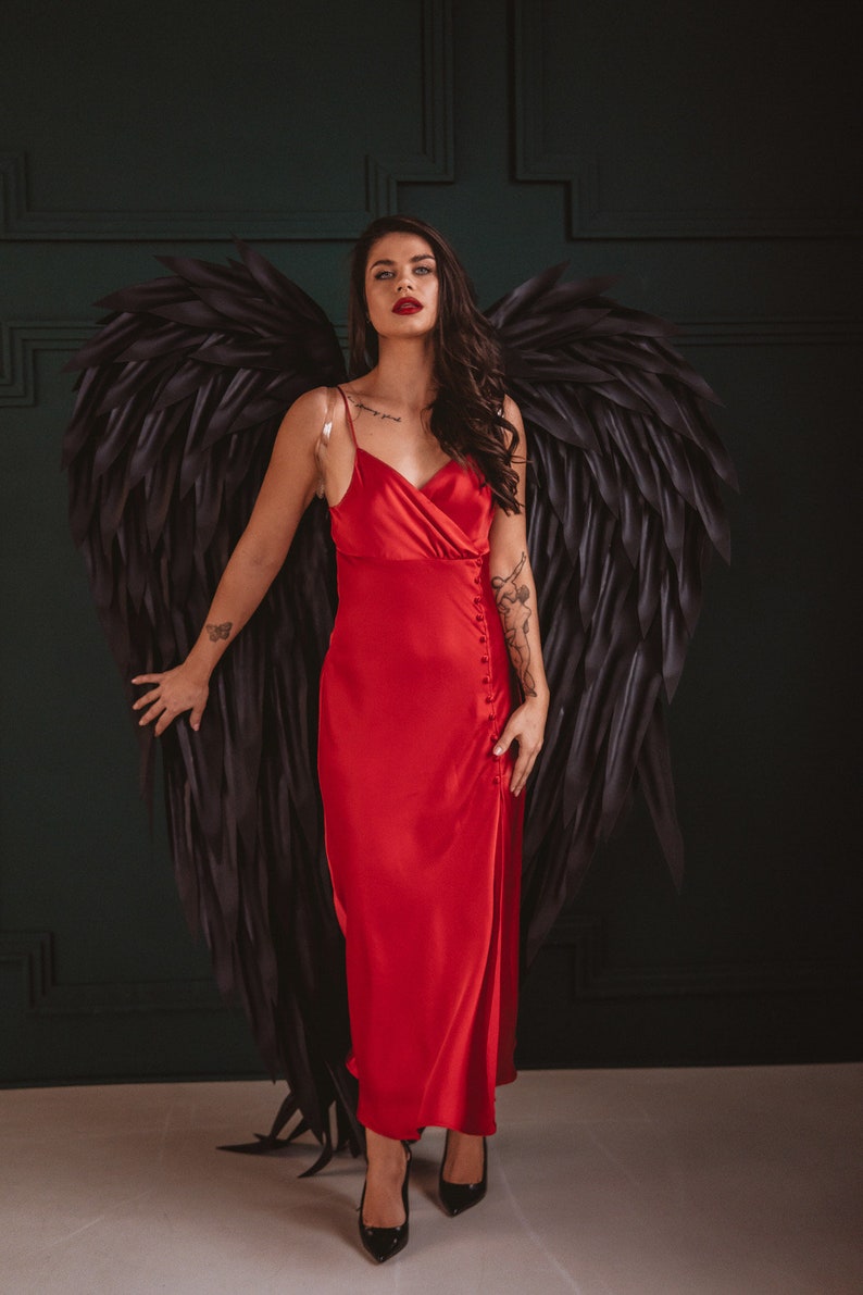 May include: A woman in a red satin dress with black angel wings. The dress has a button closure and spaghetti straps. She is wearing black heels. The background is a dark green wall.
