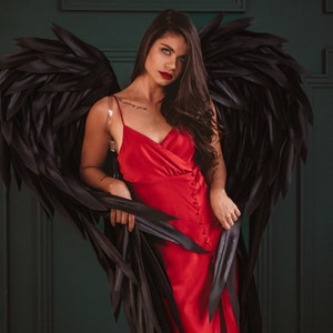 May include: A red satin dress with a button-down front and a high slit. The dress is paired with large black feathered wings. The background is a dark green wall.