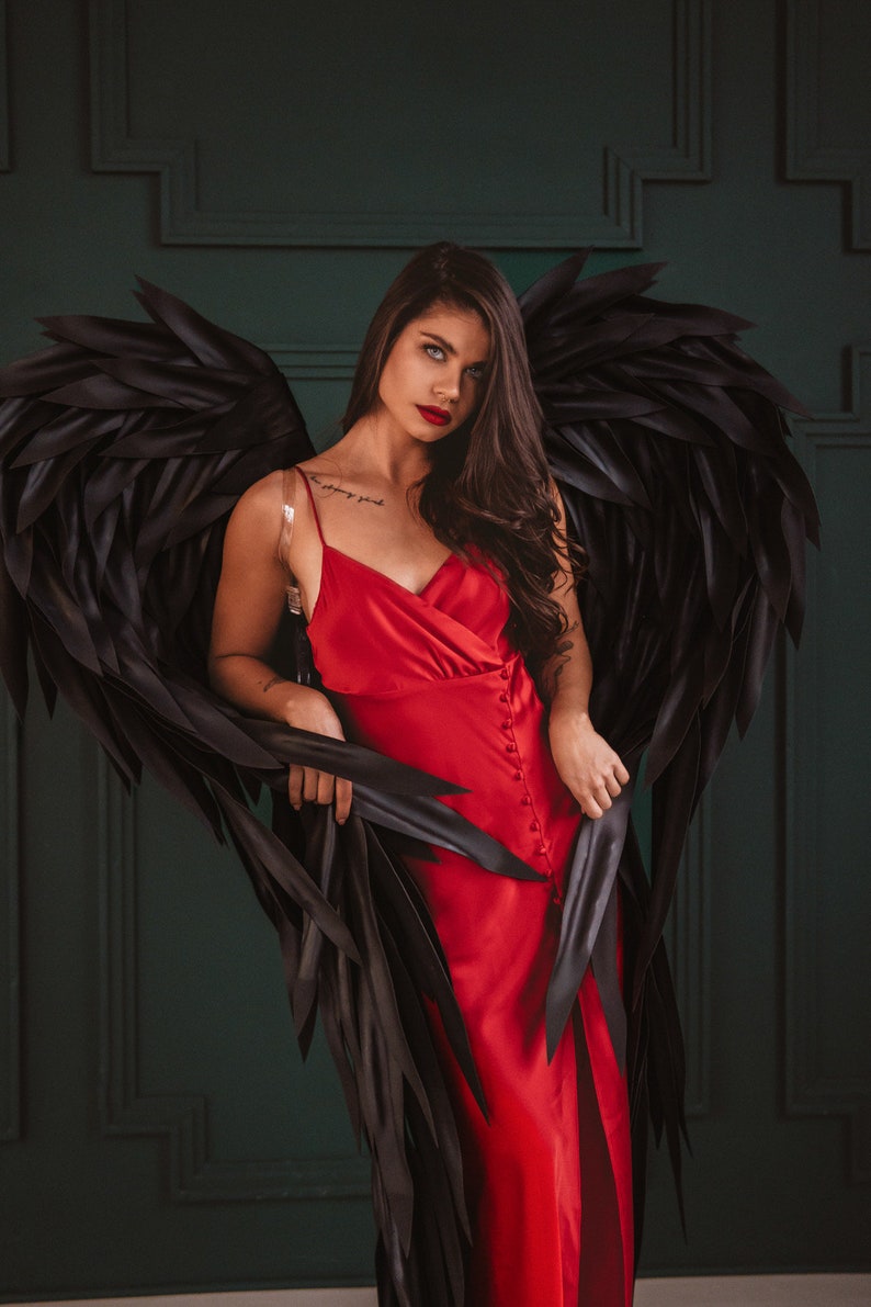 May include: A red satin dress with a button-down front and a high slit. The dress is paired with large black feathered wings. The background is a dark green wall.