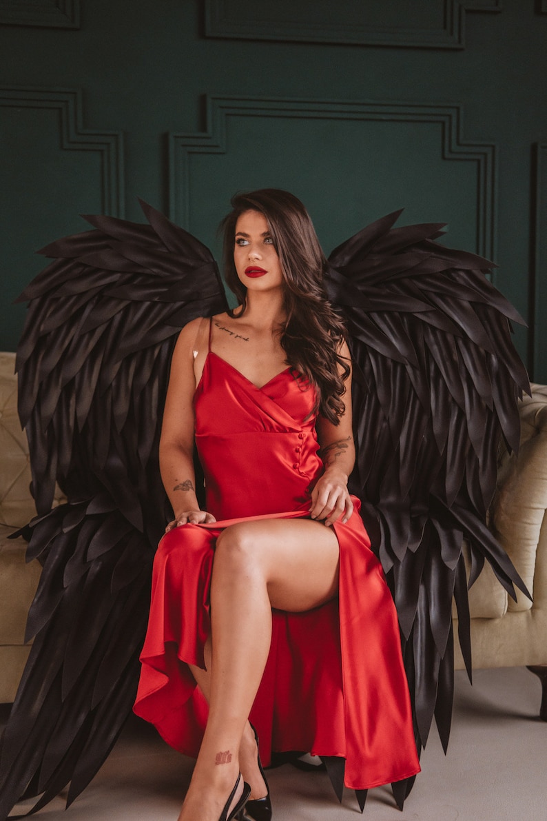 May include: A woman wearing a red satin dress and black high heels, seated on a beige sofa. Large black angel wings are behind her. The background features a dark green wall.