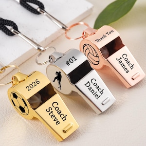 Op de afbeelding: Drie gepersonaliseerde metalen fluitjes met zwarte lanyards. Eén is goudkleurig met een voetbalafbeelding en de tekst "2026" en "Coach Steve". Een andere is zilverkleurig met een silhouet en de tekst "#01" en "Coach Daniel". De derde is roségoudkleurig met een volleybalafbeelding en de tekst "Thank You" en "Coach James".