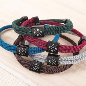 May include: A collection of rope bracelets in various colors, including blue, green, burgundy, gray, and brown. Each bracelet features a black square charm with engraved symbols and letters. The bracelets are displayed on a light wood surface.