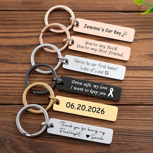 Peut inclure: Plusieurs porte-clés personnalisés avec des inscriptions gravées. Les couleurs incluent l'argent, l'or et le noir. Les messages comprennent "Jemima's Car Key" et une date, "06.20.2026". Les porte-clés sont présentés sur une surface en bois.