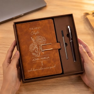 Op de afbeelding: Een beige notitieboek in een bruine doos met een gouden metalen sluiting, een pen en een vulling. De kaft is gegraveerd met bloemmotieven, de tekst "THE NEXT Chapter", de naam "JOHN AMELIA" en "Each chapter ushers in new growth."