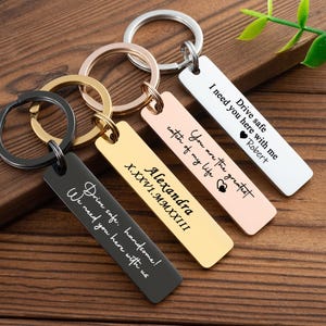 May include: A collection of personalized keychains in various colors, including black, gold, rose gold, and silver. Each rectangular keychain features engraved text, such as names and phrases like "Drive safe." The keychains are displayed on a wooden surface.