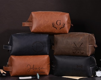 May include: Five different leather toiletry bags with personalized engravings. The bags are brown, black, and a dark brown color. The engravings include a monogram, a deer head, golf clubs, and a bow tie.