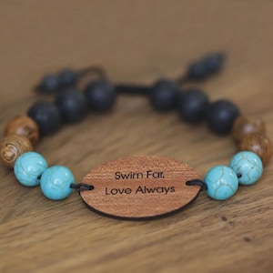 Può includere: Un braccialetto di perline con perline turchesi, di legno e nere. Un ciondolo ovale in legno al centro è inciso con le parole "Swim Far, Love Always". Il braccialetto è esposto su una superficie di legno.