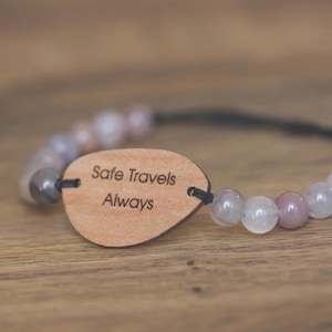 Op de afbeelding: Een armband met lichtroze, witte en grijze kralen. Het heeft een houten label met de tekst "Safe Travels Always". De armband is geregen op een zwart koord. De achtergrond is een houten oppervlak.