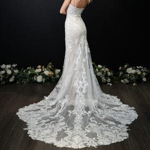 May include: A white, floor-length wedding gown with a fitted bodice and a flowing skirt. The dress features intricate lace detailing and a long train. The bride is standing with her back to the camera, with her arms crossed. The background is a dark wall with floral decorations.