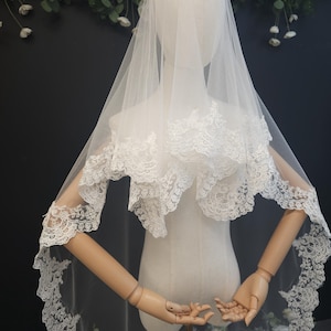 May include: A white bridal veil with intricate lace detailing. The veil is draped over a mannequin, showcasing the delicate lace trim along the edges and around the shoulders. The veil is made of a sheer, lightweight fabric.