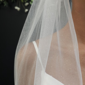 May include: A sheer, white bridal veil with a subtle glitter effect. The veil is draped over the shoulder, revealing a white dress and a glimpse of a gold earring. The fabric is delicate and airy, perfect for a wedding.