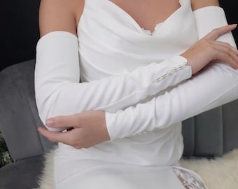 May include: A white wedding dress with a cowl neckline and spaghetti straps. The dress features long, detachable sleeves with pearl button details at the cuffs. A slit reveals a lace panel on the skirt.