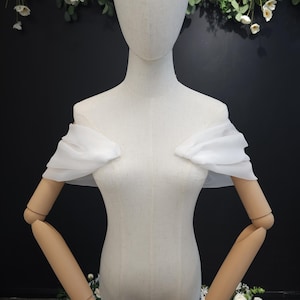 May include: A white, off-the-shoulder bridal top with ruffled sleeves is displayed on a mannequin. The mannequin is positioned in front of a dark background with floral accents. The top is made of a lightweight, flowing fabric.