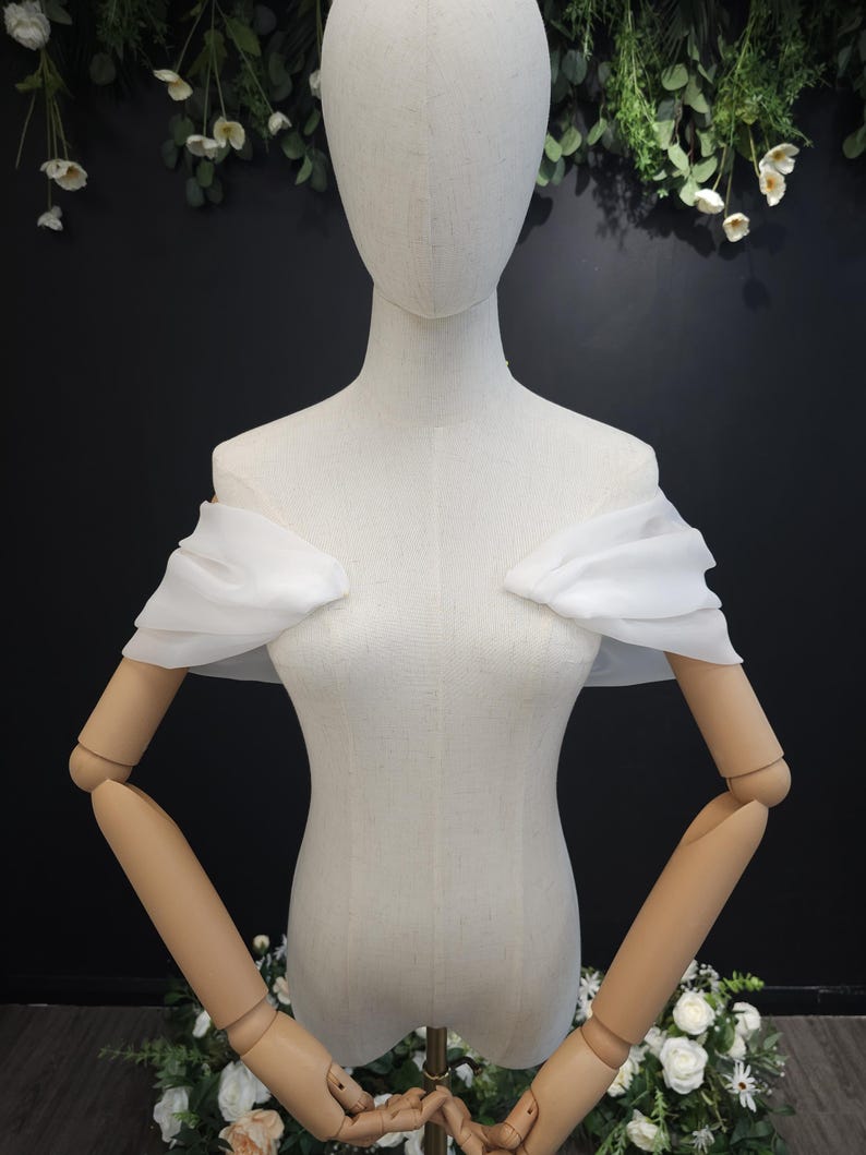 May include: A white, off-the-shoulder bridal top with ruffled sleeves is displayed on a mannequin. The mannequin is positioned in front of a dark background with floral accents. The top is made of a lightweight, flowing fabric.