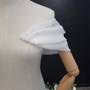 May include: A white, off-the-shoulder fabric detail draped over a mannequin's shoulder and arm. The fabric appears to be sheer and flowing, creating a soft, elegant look. The mannequin is a neutral color, and the background is dark.