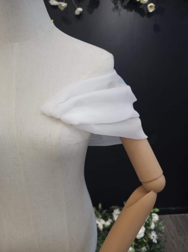 May include: A white, off-the-shoulder fabric detail draped over a mannequin's shoulder and arm. The fabric appears to be sheer and flowing, creating a soft, elegant look. The mannequin is a neutral color, and the background is dark.