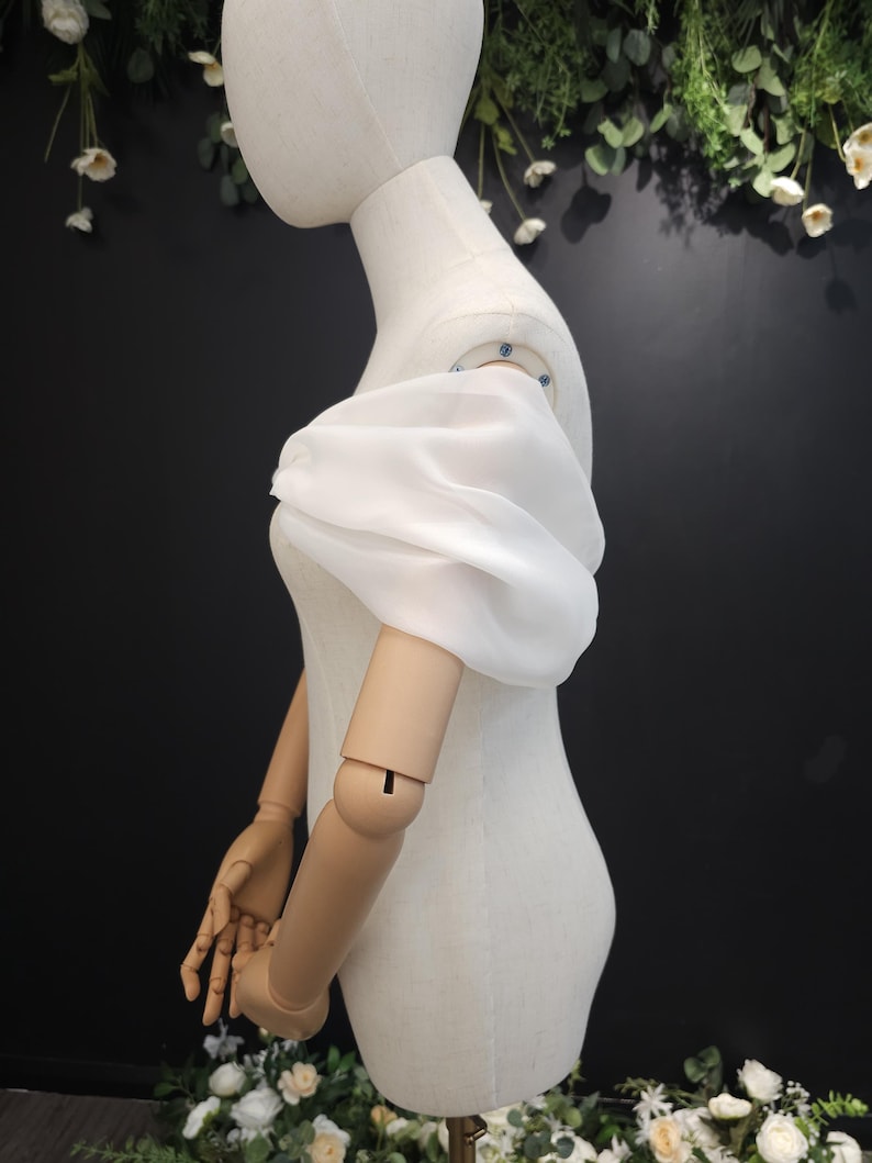 May include: A white, sheer, puff sleeve is displayed on a mannequin. The sleeve is made of a lightweight fabric and is gathered at the shoulder and wrist. The mannequin is a neutral color, and the background is dark with floral accents.
