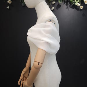 Pode incluir: Uma manga bufante branca e transparente é exibida em um manequim. A manga é feita de um tecido leve e é franzida no ombro e no pulso. O manequim é de cor neutra e o fundo é escuro com detalhes florais.