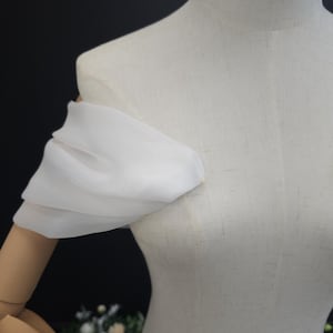 May include: A white, off-the-shoulder sleeve made of a sheer fabric, draped over a mannequin. The sleeve is gathered at the shoulder and arm, creating a soft, flowing effect. The mannequin is a light beige color.
