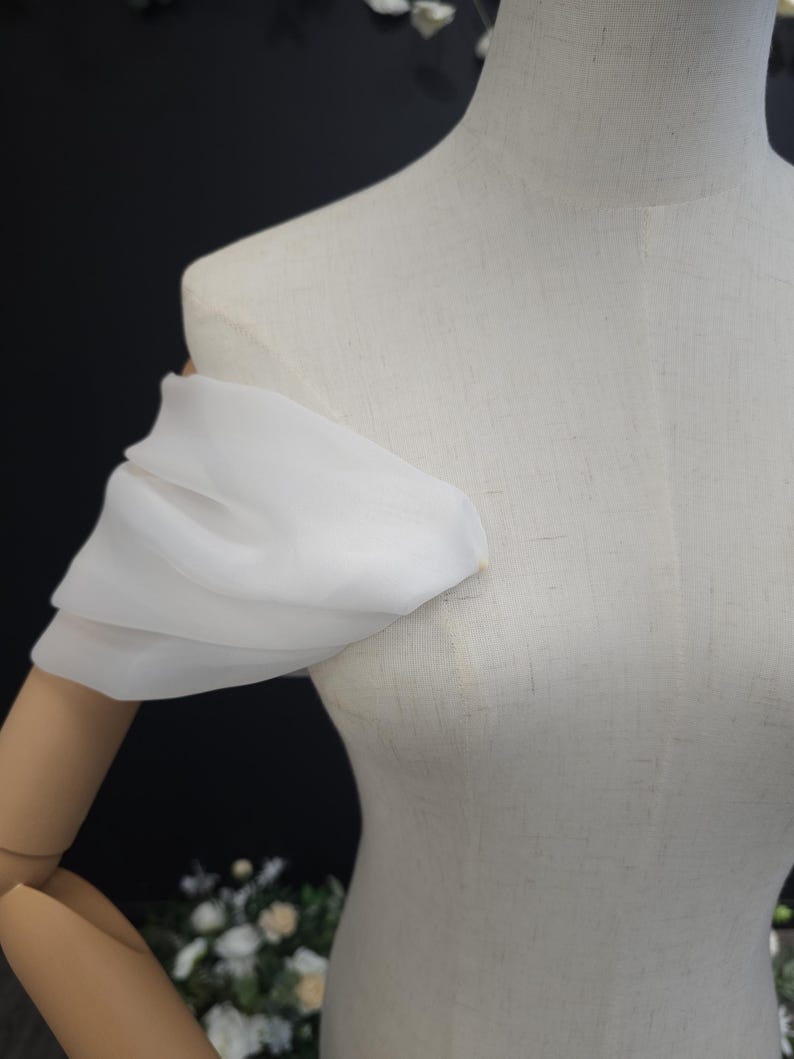 May include: A white, off-the-shoulder sleeve made of a sheer fabric, draped over a mannequin. The sleeve is gathered at the shoulder and arm, creating a soft, flowing effect. The mannequin is a light beige color.