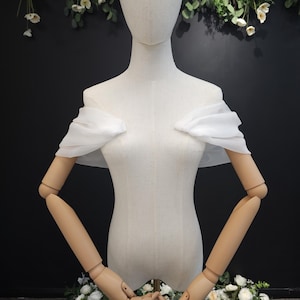 May include: A white, off-the-shoulder bridal wrap on a mannequin. The wrap is made of a sheer, flowing fabric. The mannequin has articulated wooden arms and is surrounded by floral arrangements.
