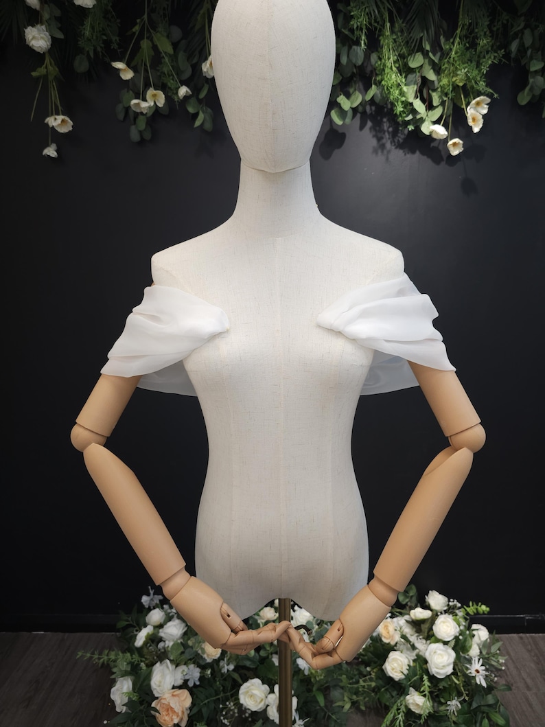 May include: A white, off-the-shoulder bridal wrap on a mannequin. The wrap is made of a sheer, flowing fabric. The mannequin has articulated wooden arms and is surrounded by floral arrangements.