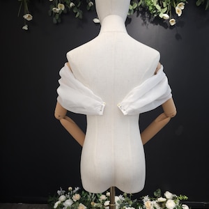 May include: A white, off-the-shoulder garment with sheer, puffed sleeves is displayed on a mannequin. The mannequin has wooden arms and is set against a dark background with floral decorations. The garment's sleeves are gathered at the wrists.