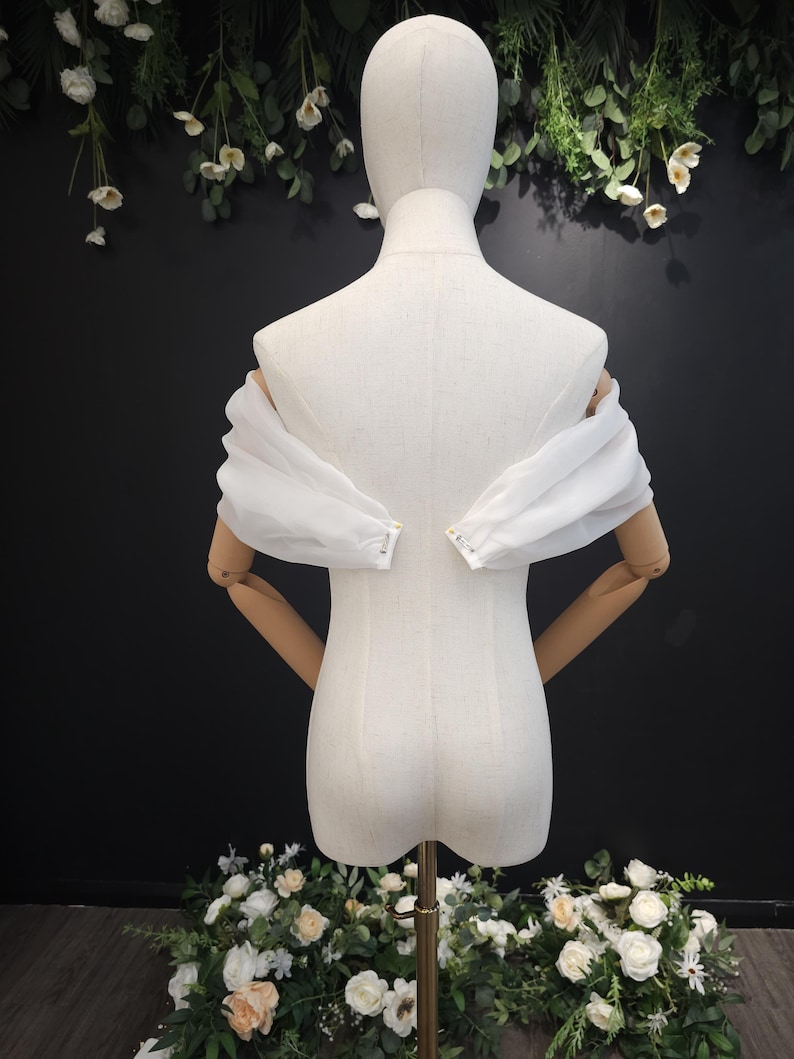 May include: A white, off-the-shoulder garment with sheer, puffed sleeves is displayed on a mannequin. The mannequin has wooden arms and is set against a dark background with floral decorations. The garment's sleeves are gathered at the wrists.