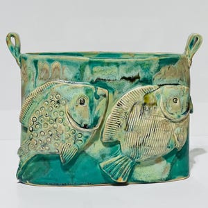 May include: A ceramic planter with two fish sculpted on the front. The planter is oval shaped and has a turquoise glaze with a green and white fish design. The planter has two handles on the top.