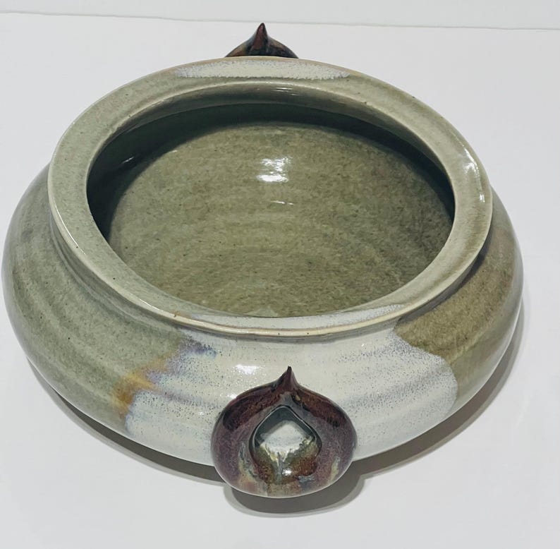 Pu&ograve; includere: Una grande ciotola in ceramica verde con tre manici marroni. La ciotola ha una glassa bianca sul fondo e sui lati. I manici hanno la forma di una lacrima e hanno una glassa marrone.
