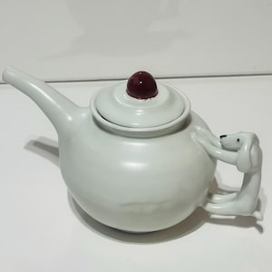 Könnte beinhalten: Eine weiße Keramik-Teekanne mit einem hundeformigen Griff und einem dunkelroten Knopf auf dem Deckel. Die Teekanne hat einen runden Körper und einen langen Ausguss. Der Hundegriff ist weiß.