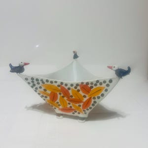 Può includere: Una ciotola in ceramica bianca con bordo appuntito a quattro angoli, caratterizzata da tre piccole statuette di pulcinella di mare appollaiate sui bordi. L'esterno è decorato con un motivo di foglie arancioni e gialle e piccoli punti grigi, su quattro piedini.
