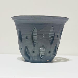 May include: A dark gray ceramic orchid pot with a flared rim and decorative cut-out leaf-shaped patterns. The pot has a series of holes for drainage and ventilation. The pot is approximately 6 inches in diameter and 5 inches tall.
