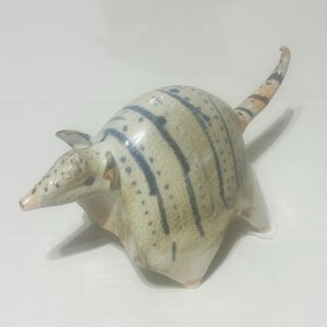 May include: A ceramic armadillo figurine featuring a cream-colored body with dark blue vertical stripes and speckled markings. The sculpture has a pointed snout, small ears, and a long, slender tail with alternating dark and light bands.