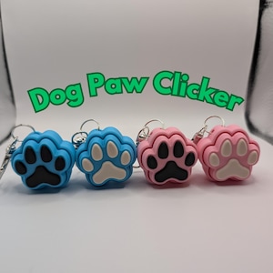 May include: Four dog paw clickers in different colors. Two are blue with black paw prints, one is white with a black paw print, and one is pink with a white paw print.