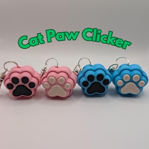 May include: Four cat paw clickers in pink, white, blue, and white. The clickers are shaped like a paw print and have a silver ring.
