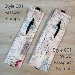Peut inclure: Deux étuis à stylos rectangulaires en tissu avec un fond crème et des motifs de tampons de passeport vintage en encre violette, bleue et rouge. Chaque étui contient un stylo noir et possède une petite languette noire. Texte Style 001 Passport Stamps.