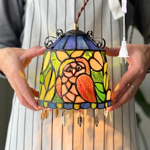 May include: Stained glass pendant lamp featuring a colorful owl design. The lamp has a blue, yellow, and orange color scheme with decorative metal feather accents. The lamp is suspended by a brown cord and has a white power cord.