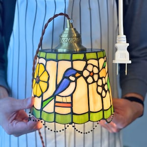 May include: A stained glass pendant lamp with a brass fixture and a white electrical plug. The lamp shade features a blue bird, yellow flowers, and white flowers with green and yellow accents. The shade is approximately 6 inches in diameter.