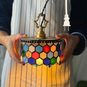 May include: A stained glass pendant lamp with a vibrant hexagonal design. The lamp features a mix of colours including blue, red, yellow, and green. It has a brass-coloured top and a decorative hanging element. The lamp is designed to be plugged into a UK outlet.