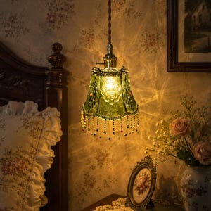 May include: A green glass pendant lamp with a brass fitting and beaded fringe, hanging in a bedroom. The lamp has a floral design on the shade. The room features floral wallpaper and a framed picture, creating a vintage aesthetic.