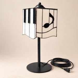 May include: A table lamp featuring a square stained glass shade with a black and white piano key pattern on one side and a black musical note design on the other. The lamp has a black metal base, a thin vertical stand, and a black power cord.