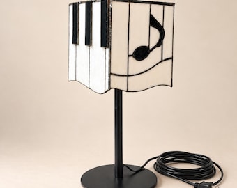 Puede incluir: Una lámpara de mesa con una pantalla cuadrada de vidrio tintado que presenta un patrón de teclas de piano en blanco y negro en un lado y una nota musical negra en el otro. La lámpara tiene una base de metal negro, un soporte fino y un cable negro.