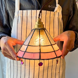 May include: A stained glass pendant lamp with a brass fixture and a pink, peach, and clear glass shade. The shade has a geometric design and is adorned with purple crystal dangles. The lamp is held by a person wearing a striped apron.